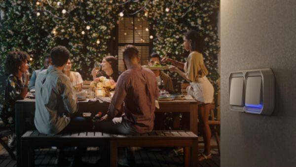 Un groupe de personnes est assis le soir autour d'une table à l'extérieur, éclairé par des guirlandes lumineuses, tandis qu'une personne propose de la nourriture.