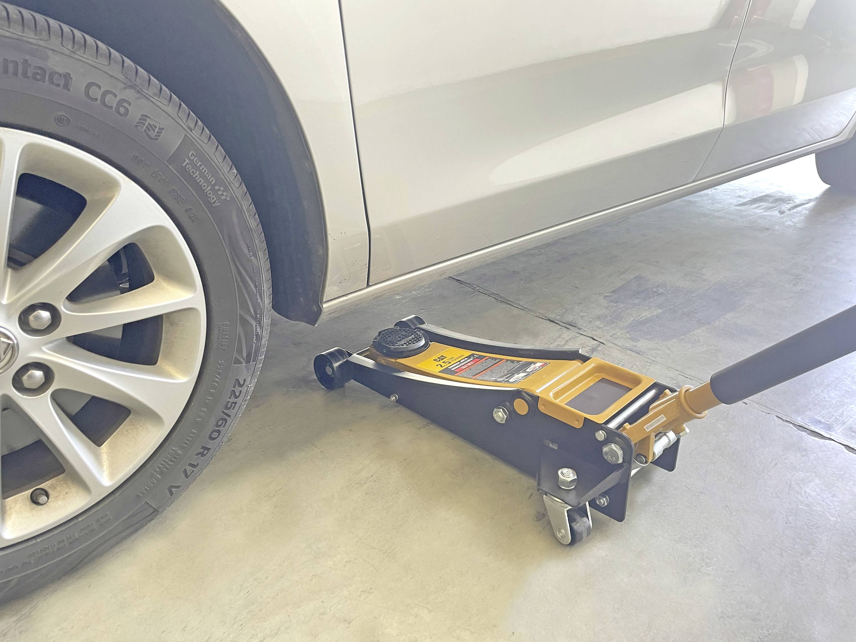 'Un cric soulevant une voiture argentée dans un garage. Le cric, placé sous le châssis de la voiture, soulève la roue avant gauche du sol.'