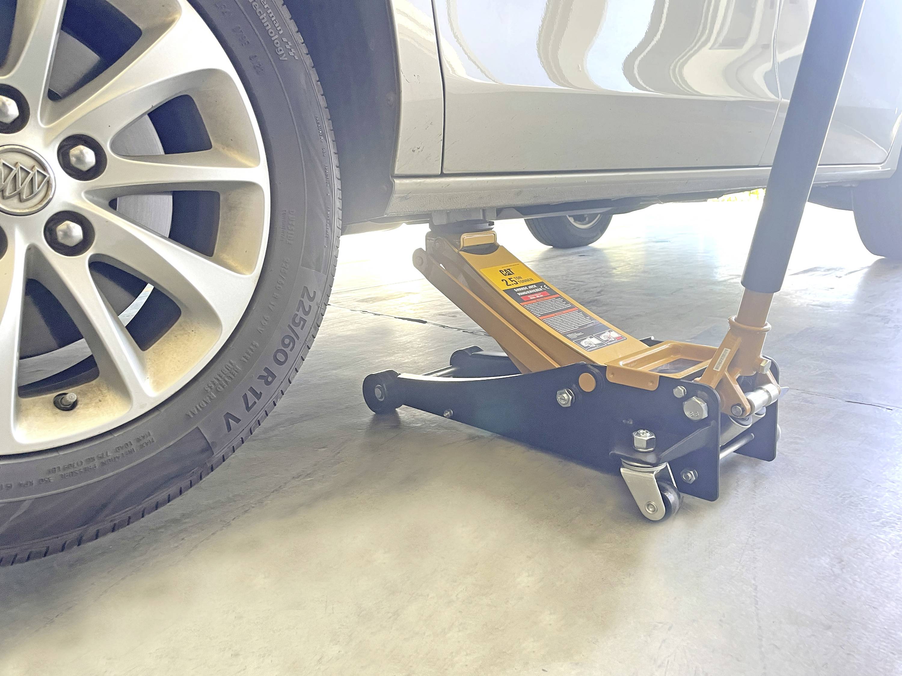 Une voiture est soulevée avec un cric hydraulique sous le pneu avant gauche dans un garage. La roue de la voiture et une partie de sa carrosserie sont visibles.