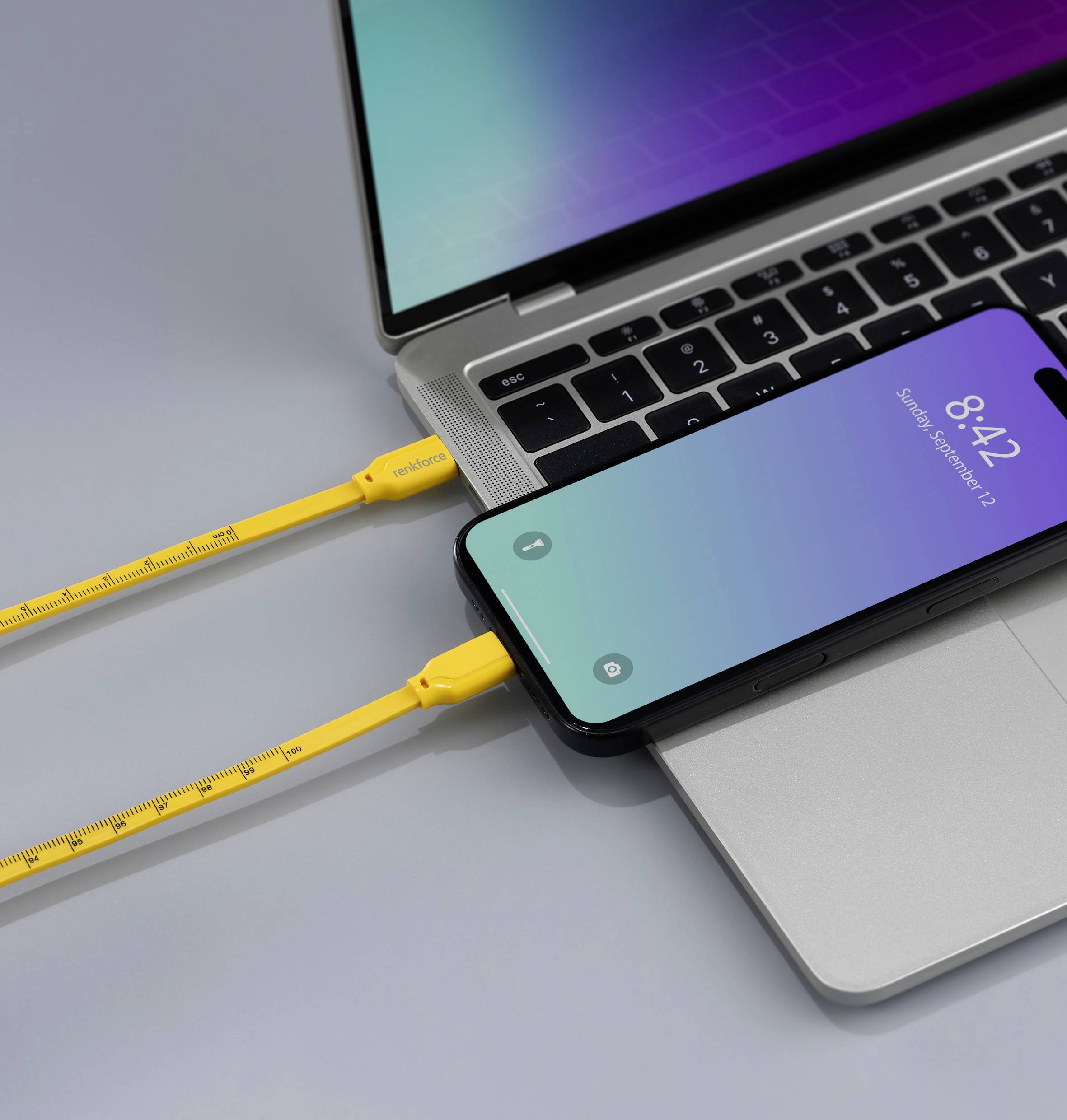 Un smartphone est posé sur un ordinateur portable et se charge via un câble jaune. L'ordinateur portable a un boîtier argenté, et l'écran du smartphone affiche l'heure 8:42.