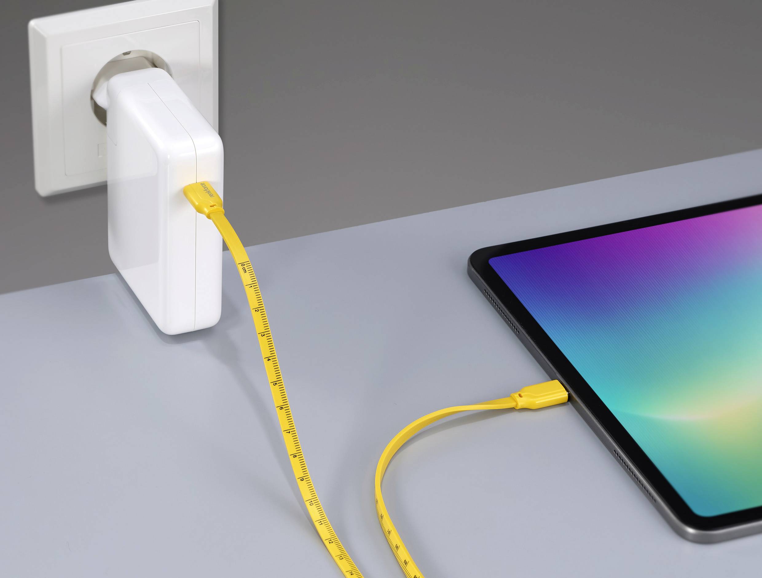 Un câble USB jaune relie un chargeur branché à une prise électrique à une tablette posée sur une table grise.
