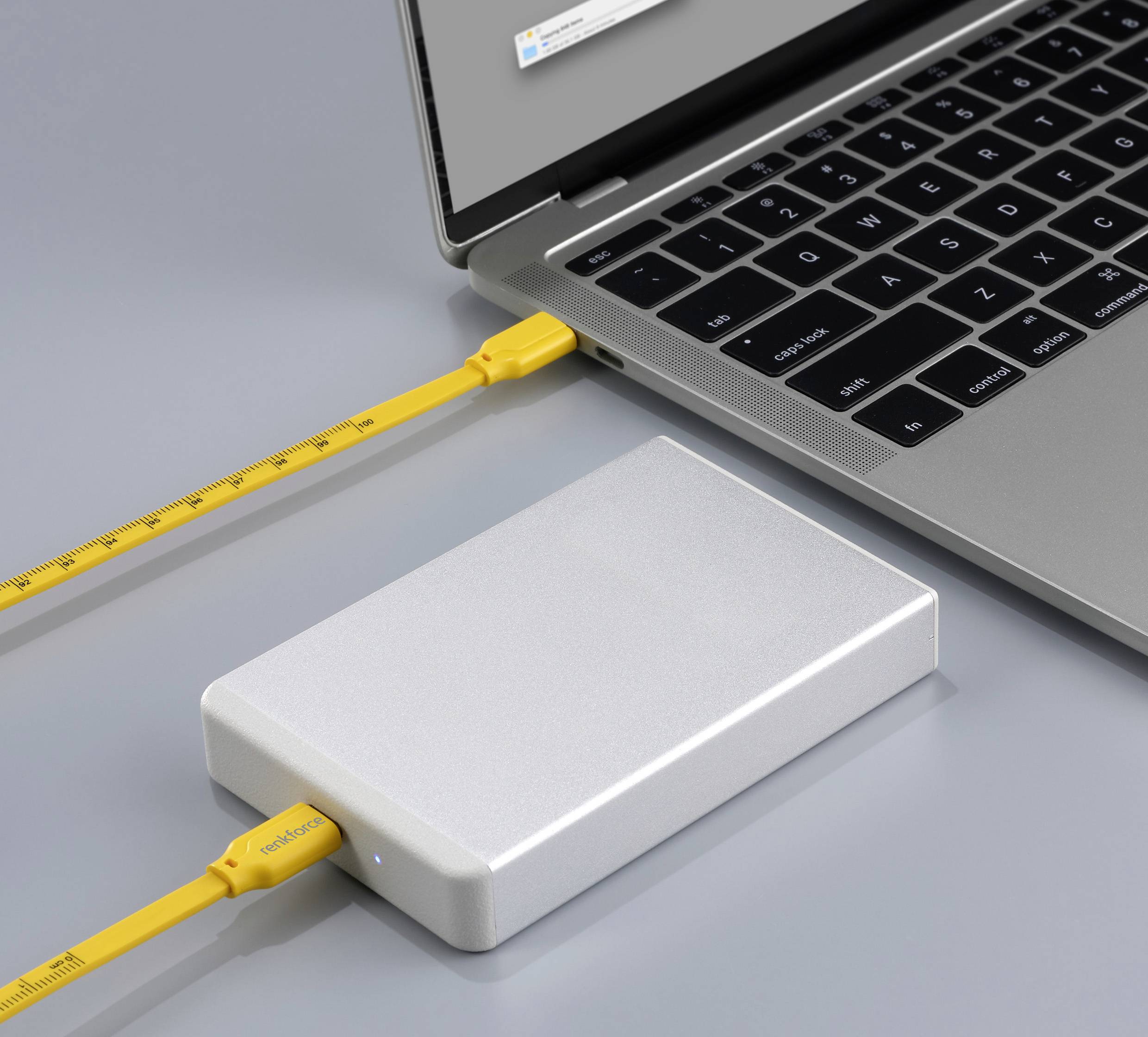 Un disque dur externe argenté est connecté à un ordinateur portable par un câble USB jaune, affichant un écran gris.