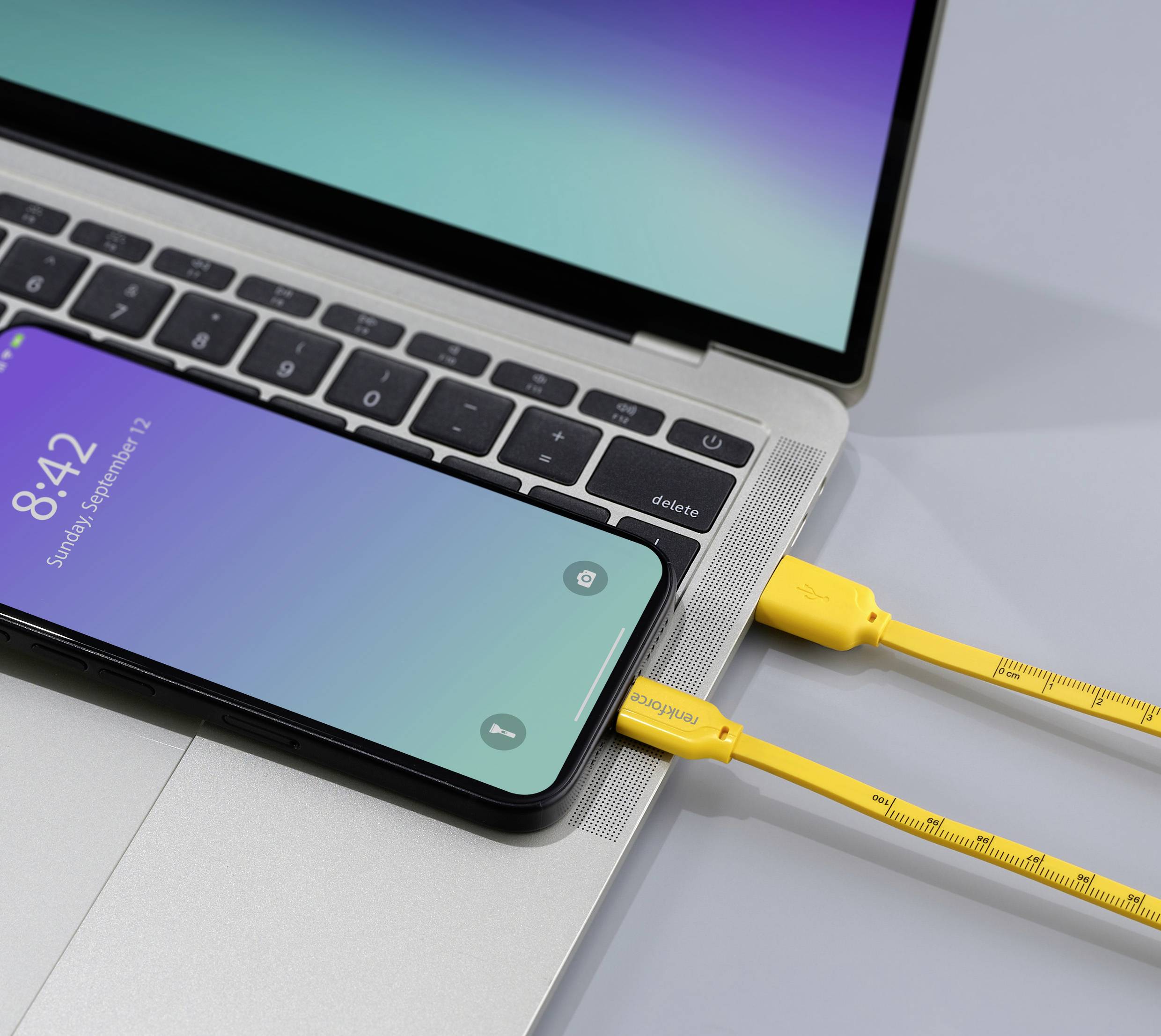 Un smartphone relié à un ordinateur portable par un câble USB jaune est posé sur une table. L'écran du téléphone affiche l'heure 8:42.