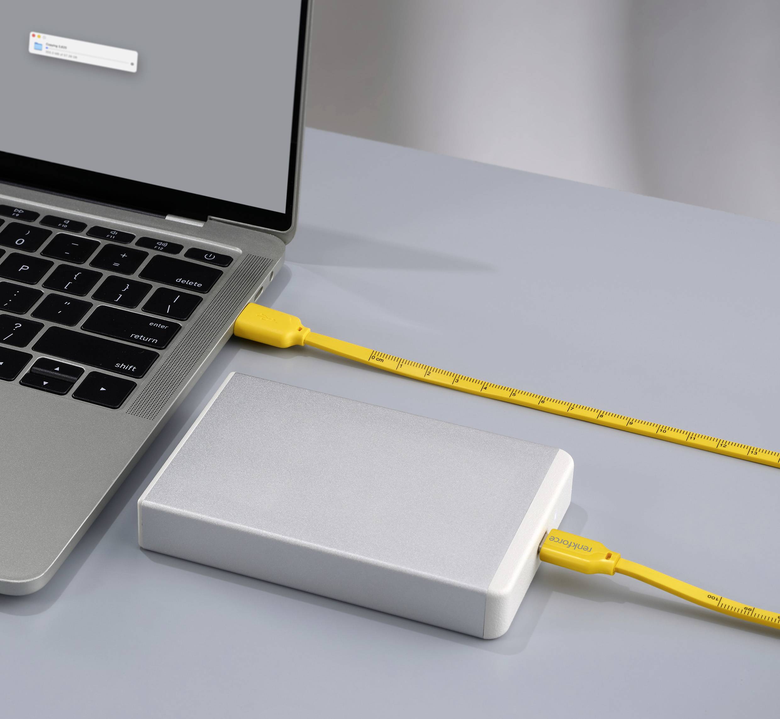 Un ordinateur portable avec un écran gris, connecté à un disque dur externe. Le câble jaune présente des marquages semblables à un mètre ruban.