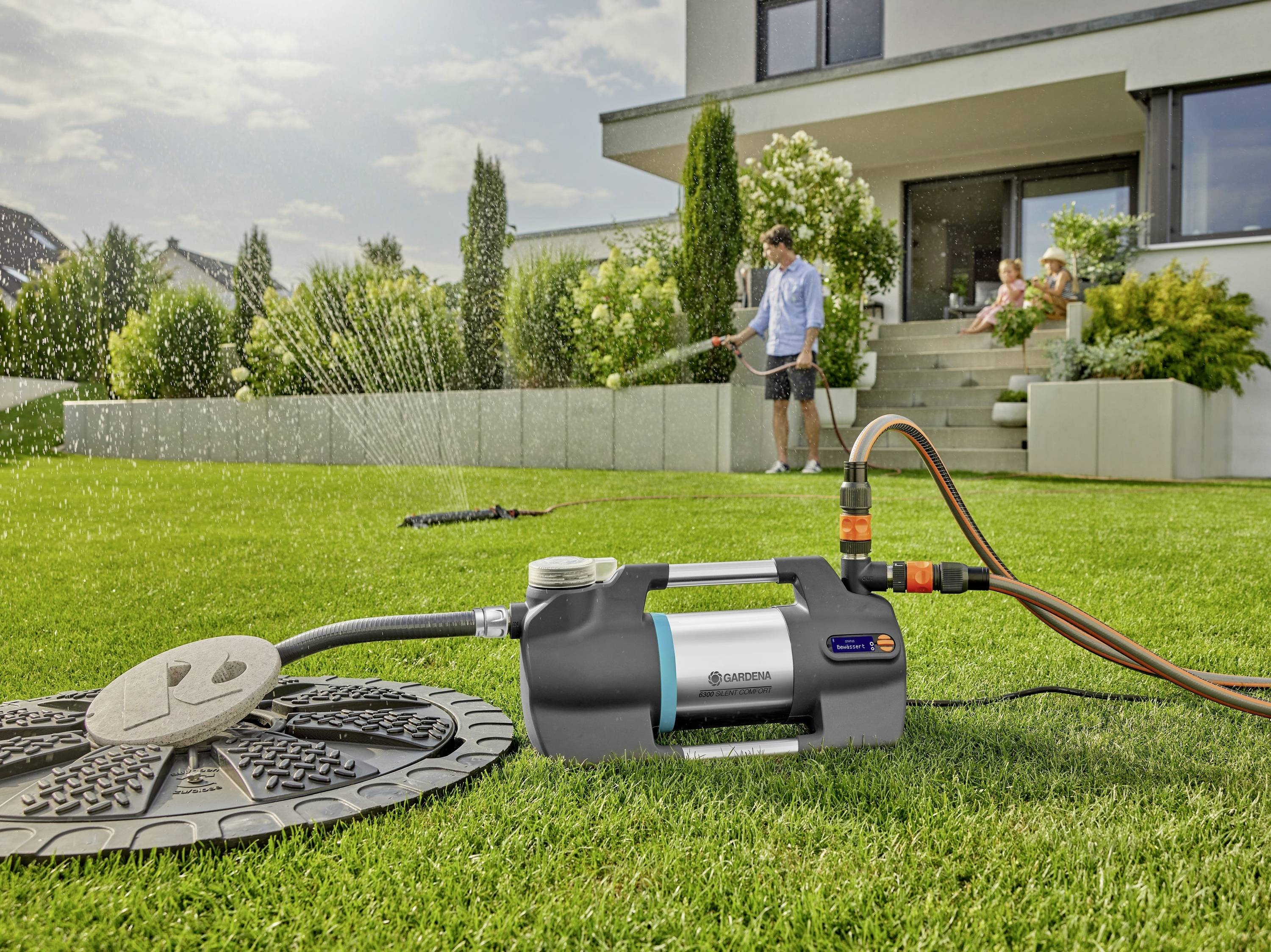 Un homme arrose la pelouse d'un jardin moderne à l'aide d'un système d'irrigation, pendant que deux personnes sont assises sur la terrasse.