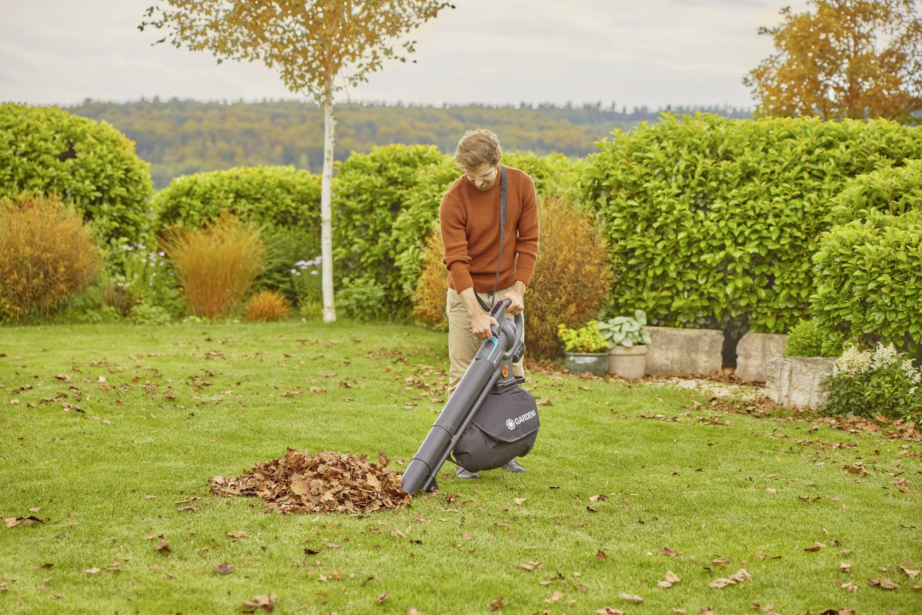 Un homme sur une pelouse utilise un aspirateur à feuilles pour enlever un tas de feuilles. L'arrière-plan montre des haies vertes et des arbres automnaux.