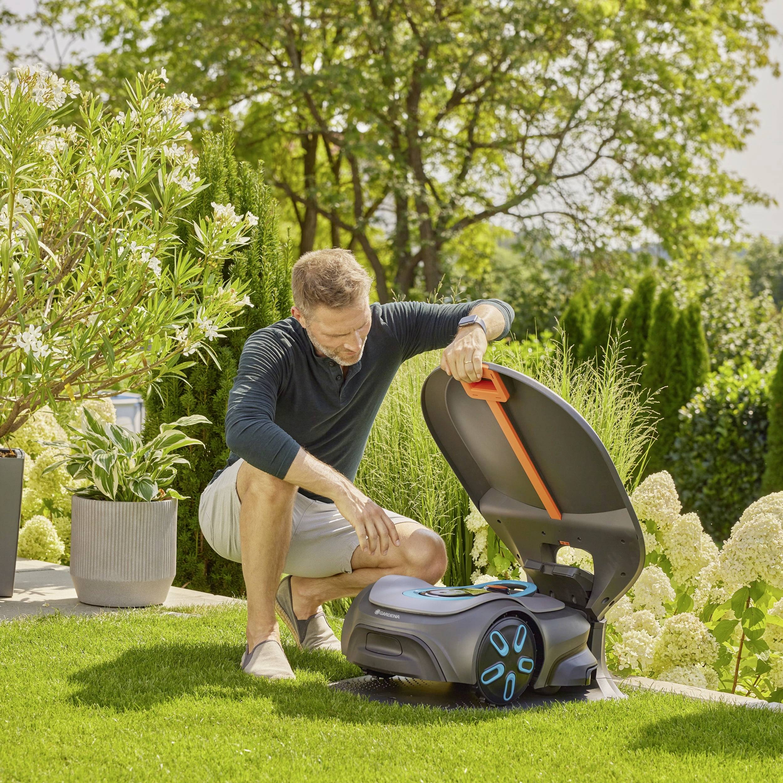 Un homme dans un jardin ouvre le couvercle d'un robot tondeuse. Entouré de plantes et d'arbres par temps ensoleillé.