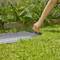 Une personne touche un tapis de sol GARDENA bleu dans un jardin, entourée de plantes vertes et de fleurs mauves, par une journée ensoleillée.