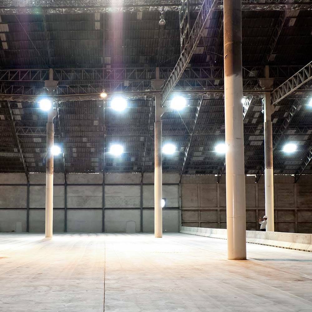Grande halle d'entrepôt vide avec des plafonds hauts, éclairée par des lumières vives suspendues au plafond. Une personne se tient près d'un mur au fond de l'espace.