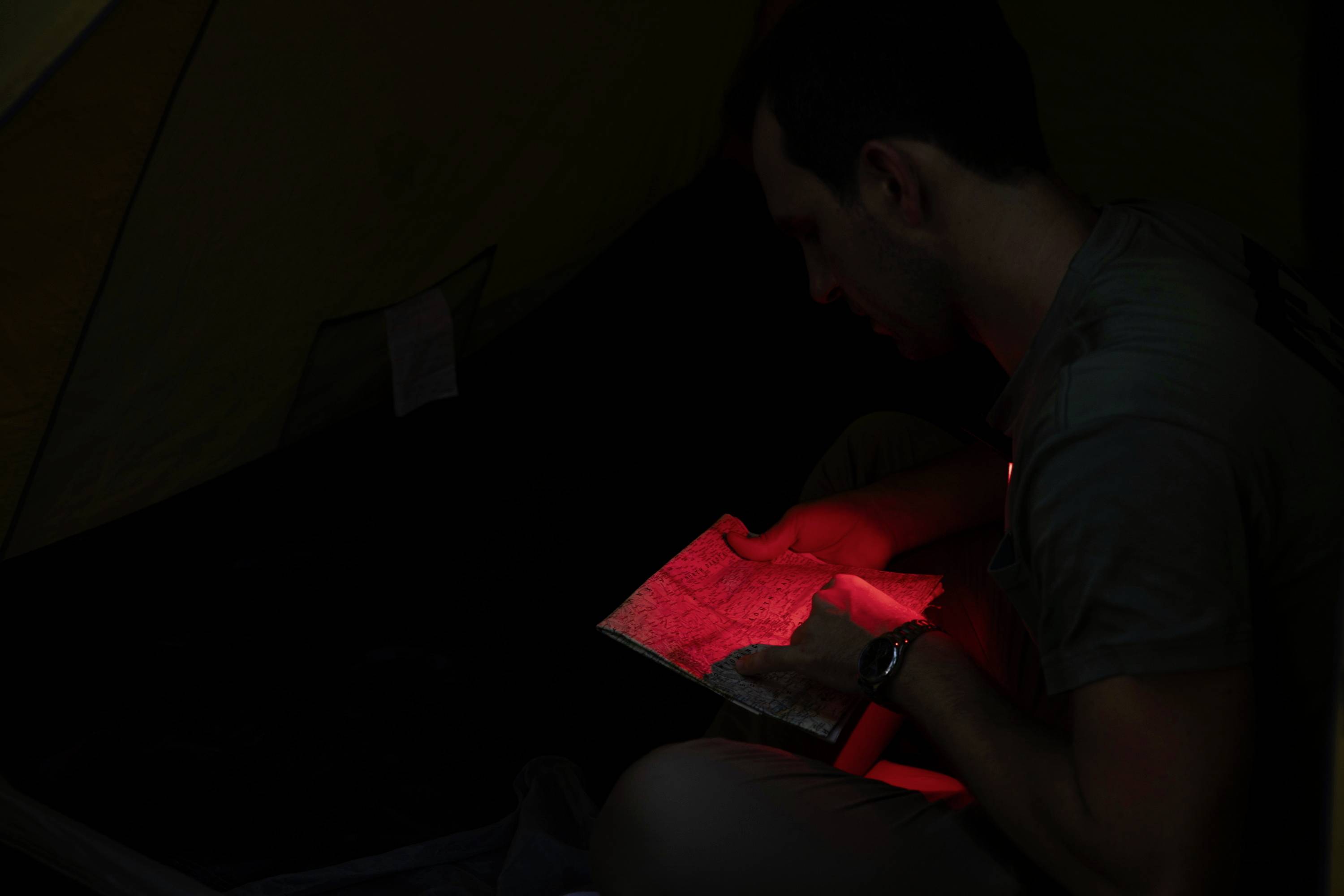 Une personne lit une carte à l'aide d'une lampe de poche rouge à l'intérieur d'une tente. Environnement sombre avec un focus sur la carte illuminée.