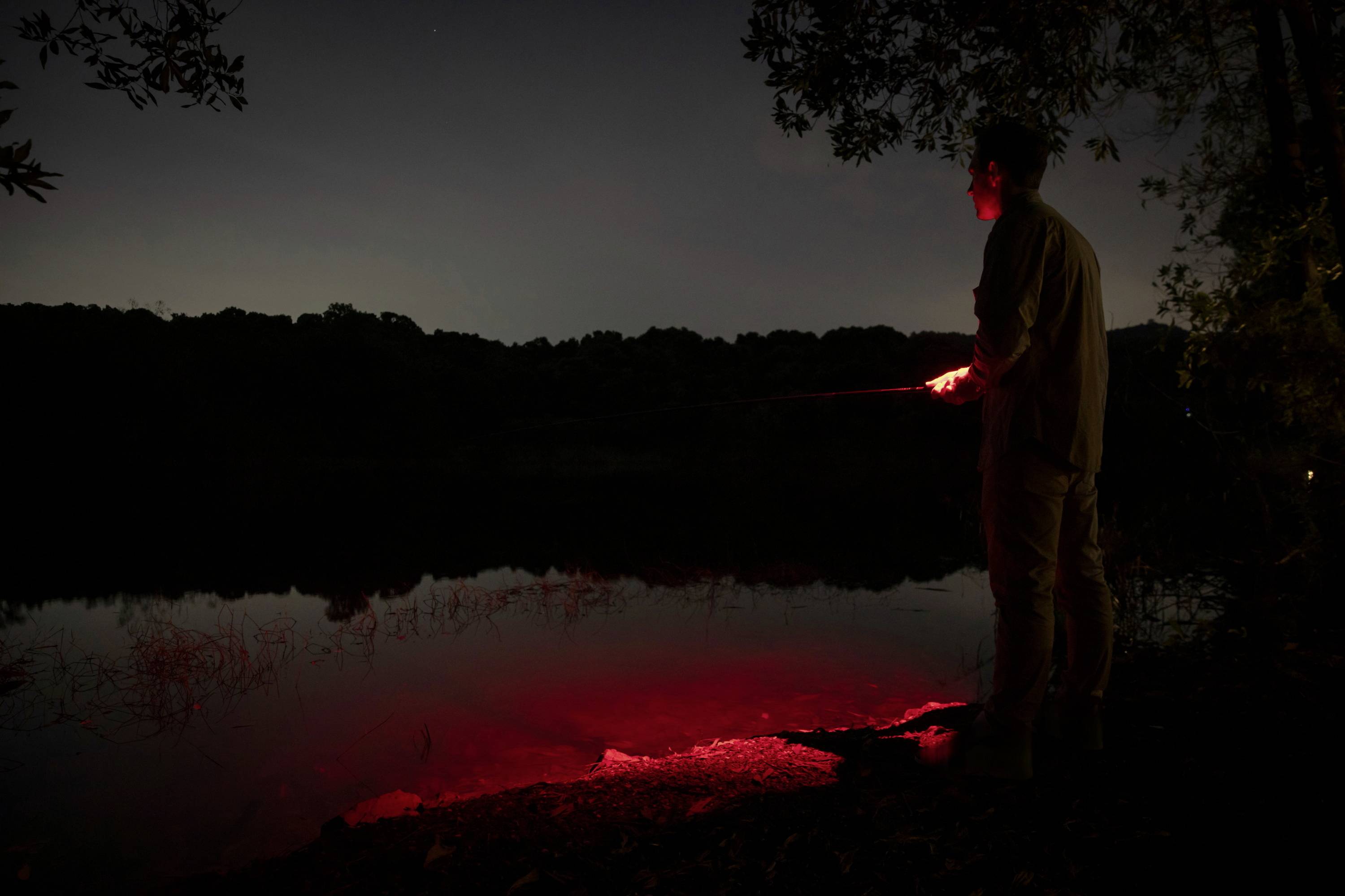 Une personne pêche au bord de la rivière la nuit, éclairée par une lumière rouge. Des arbres et de l'eau se reflètent en arrière-plan.