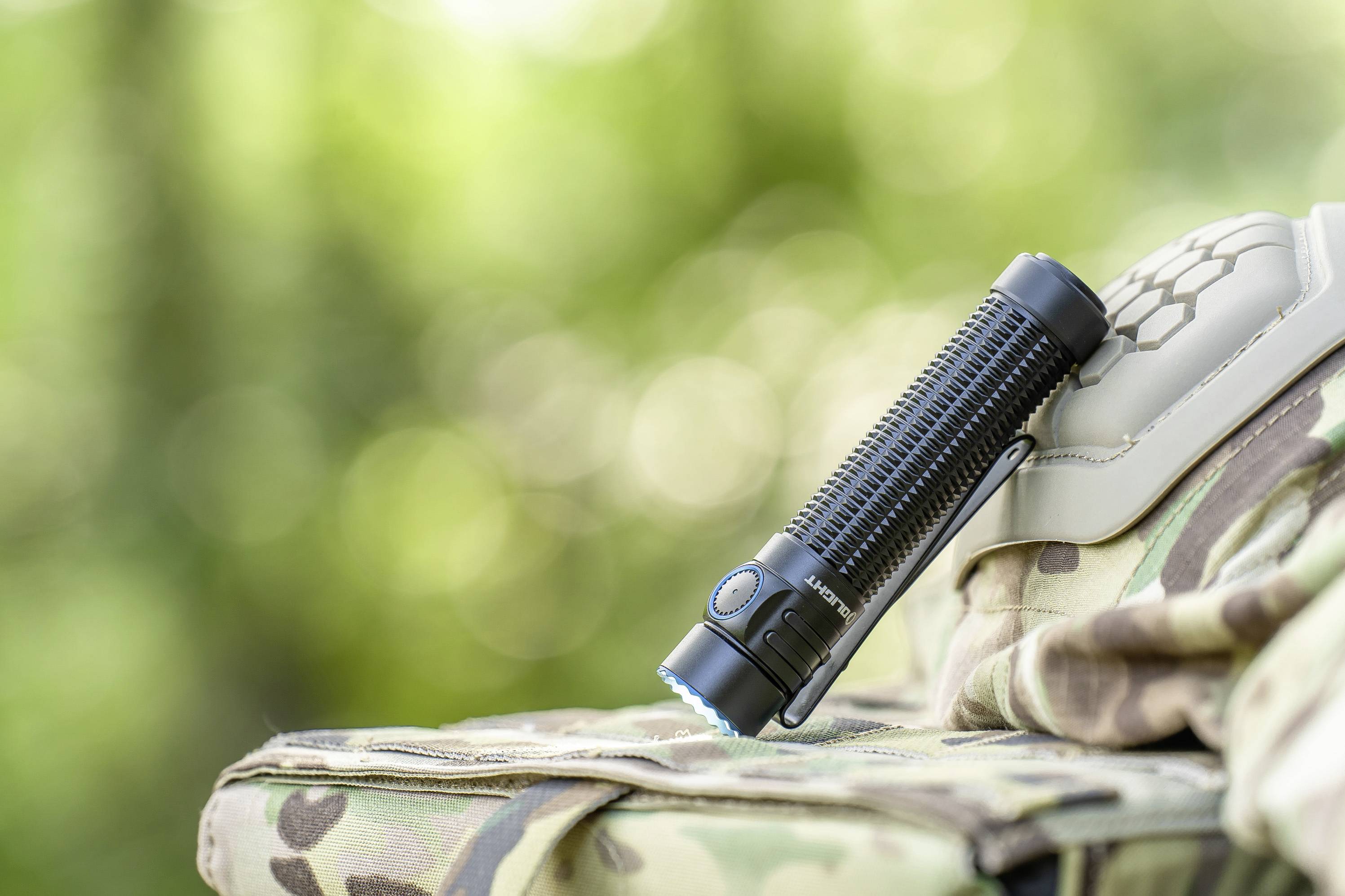 Une lampe de poche au boîtier noir repose sur un sac camouflé devant un arrière-plan de feuillage vert flou.