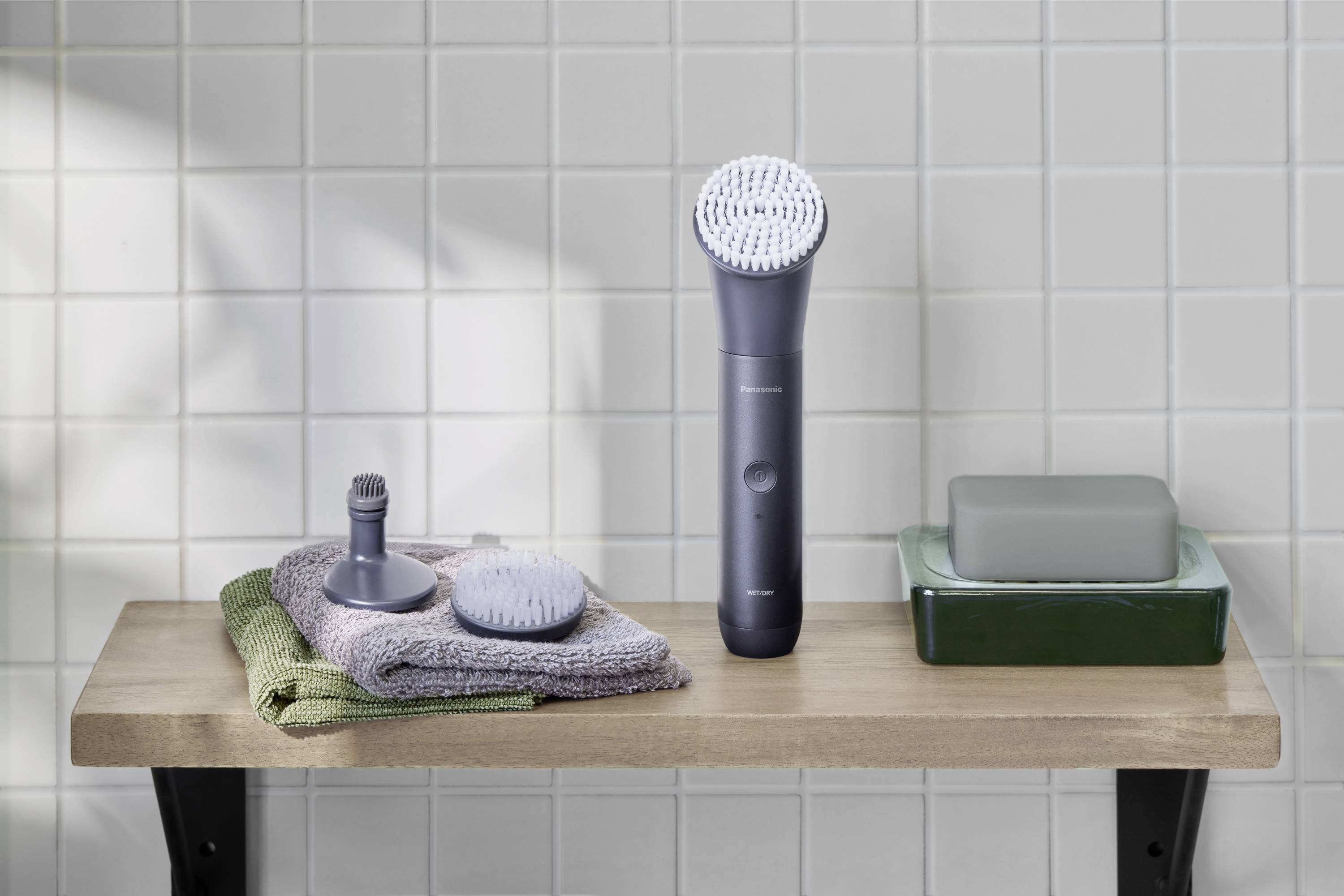 Brosse électrique de nettoyage du visage avec accessoires et porte-savon sur une étagère en bois, devant un mur carrelé blanc dans la salle de bain.