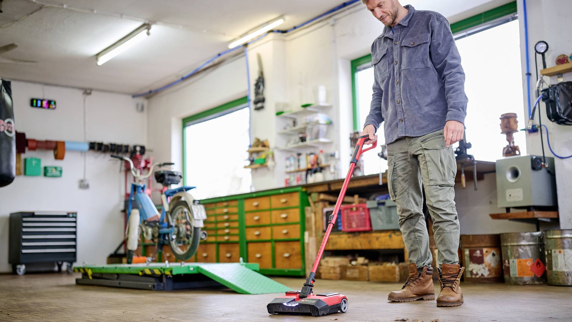 Une personne aspire le sol dans l'atelier. À l'arrière-plan, un vélo est placé sur un pont élévateur. Des outils et des tiroirs sont visibles.