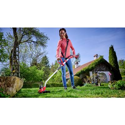 Une femme tond la pelouse avec un coupe-herbe rouge dans un jardin devant une maison au toit rouge. Des arbres se trouvent en arrière-plan.