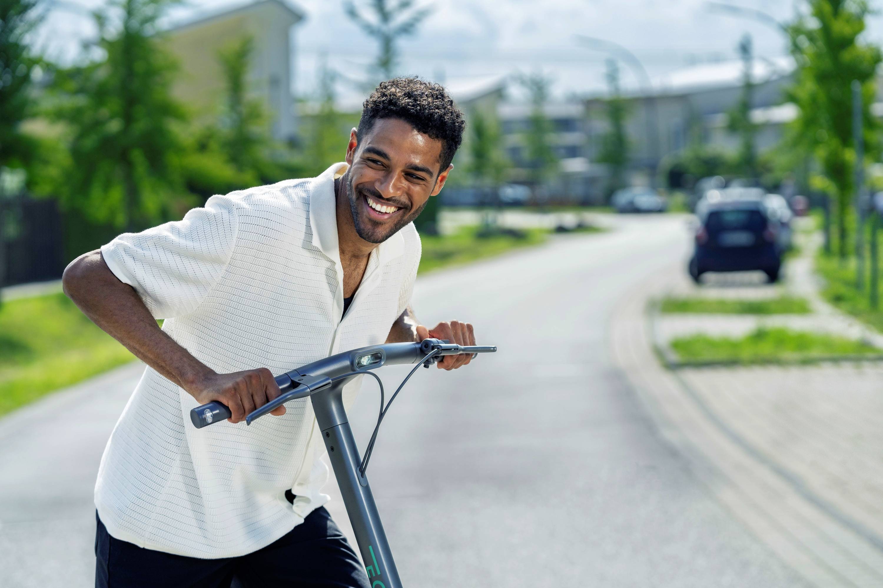 Une personne souriante conduit une trottinette électrique le long d'une rue calme bordée d'arbres, en pleine journée.