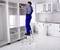 Un homme en tenue de travail est debout sur une échelle et installe un meuble dans une cuisine blanche.