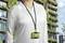 Une femme portant un t-shirt blanc porte un badge vert suspendu à un cordon noir. Un bâtiment verdoyant est visible en arrière-plan.
