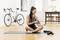 Une femme est assise sur un tapis de yoga, utilisant un pistolet de massage sur son bras. Un vélo se trouve en arrière-plan, des haltères sont posées sur le sol.