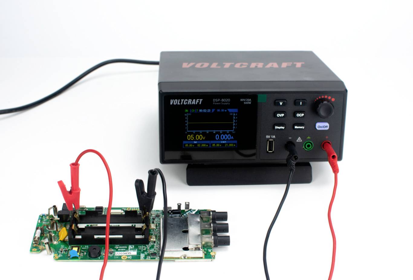 Un bloc d'alimentation numérique avec affichage LCD montre les valeurs de tension. Deux câbles sont connectés à une carte de circuit imprimé verte posée sur une table.