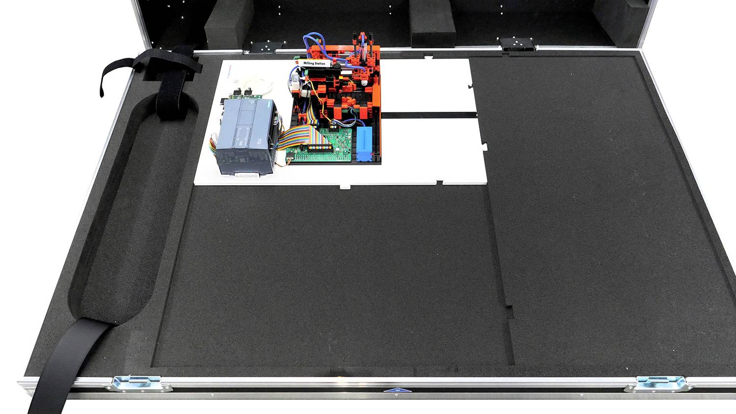 Une boîte métallique ouverte contenant un circuit électronique et des câbles sur un support en mousse ; à gauche, un compartiment avec une sangle.