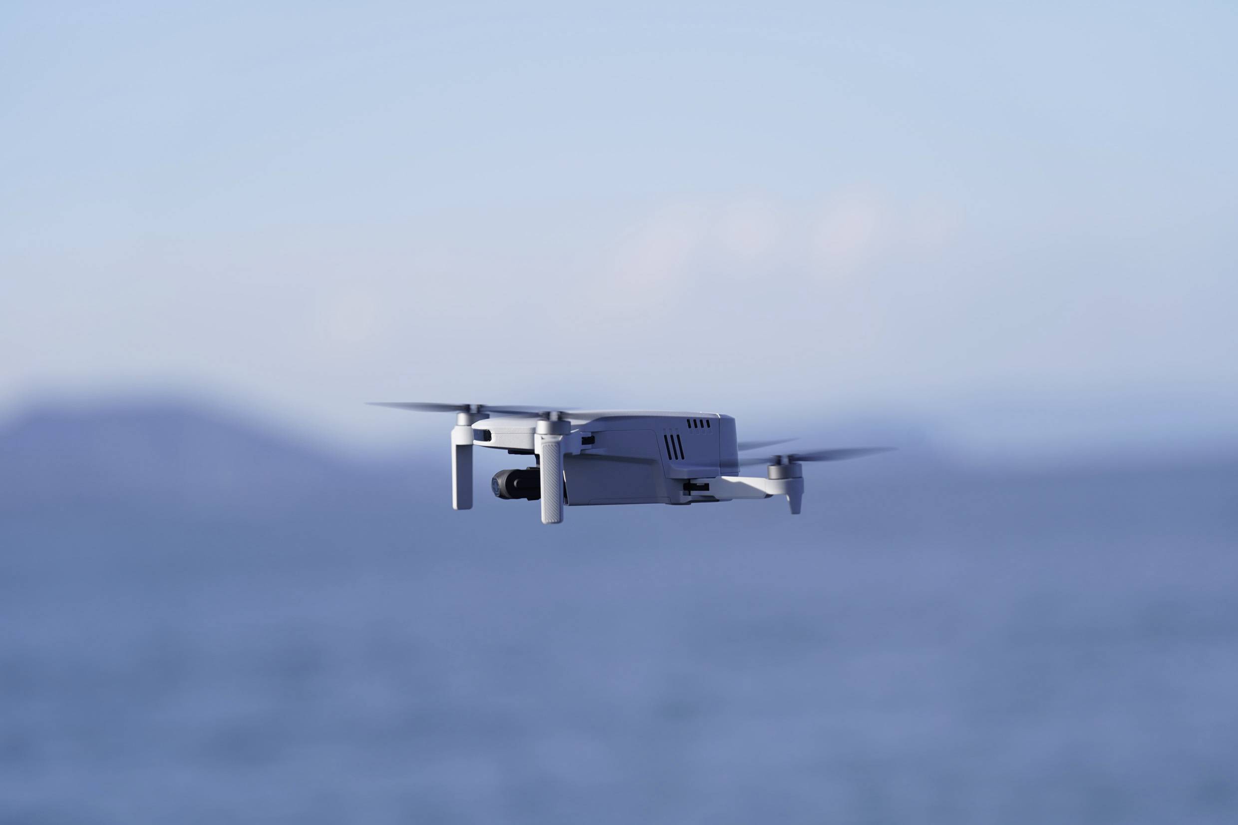 Un drone blanc plane au-dessus de la mer bleue. Des collines floues sont visibles en arrière-plan.