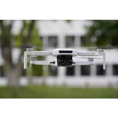 Un drone blanc plane dans les airs devant un arrière-plan flou composé d'arbres et d'un bâtiment à plusieurs étages.