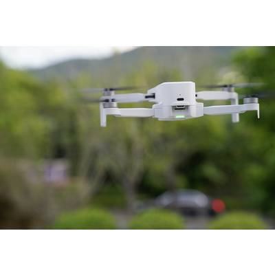 Un drone blanc plane à l'extérieur devant un paysage vert flou. Il a quatre rotors et une lumière verte brillante à l'avant.