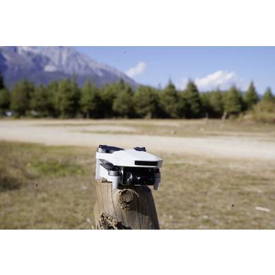 Un petit drone blanc est posé sur un poteau en bois dans un paysage forestier avec des montagnes en arrière-plan sous un ciel bleu.