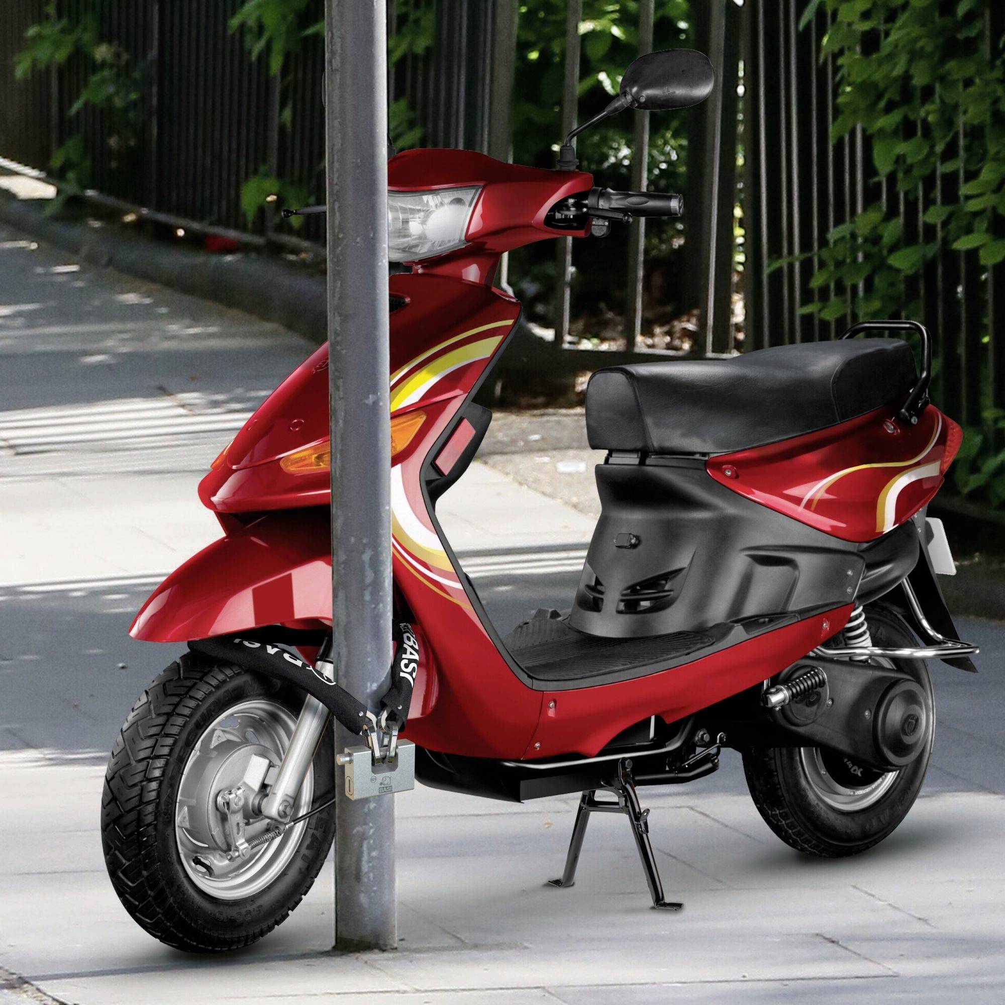 Un scooter rouge est appuyé contre un grand poteau métallique sur un trottoir, à côté d'une clôture avec des feuilles vertes en arrière-plan.