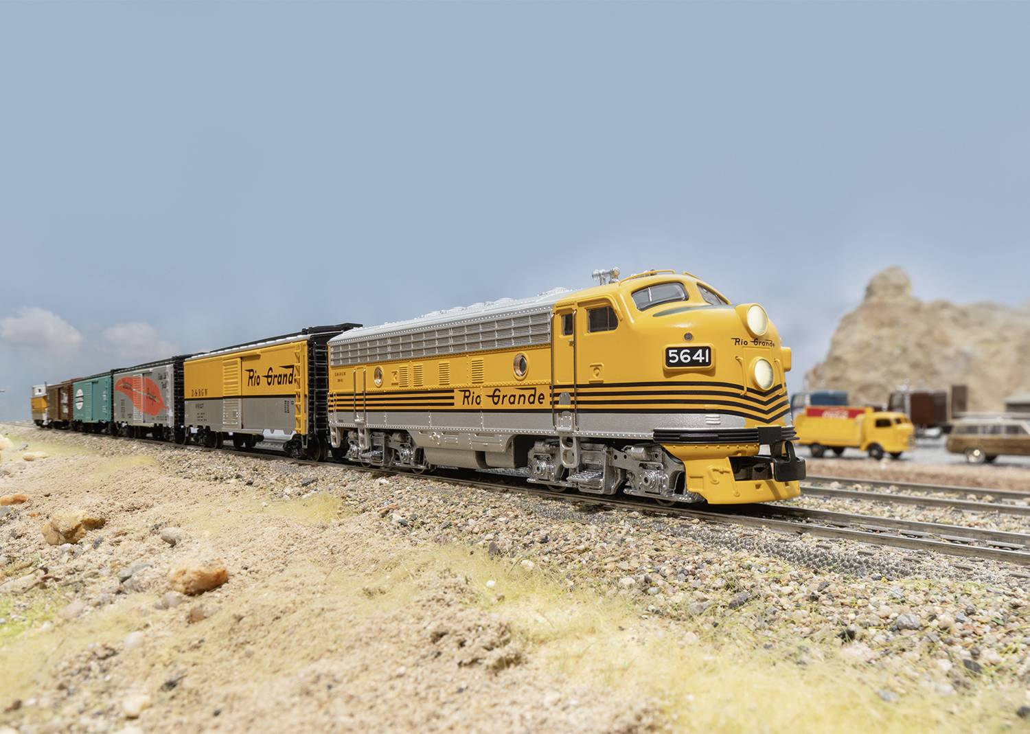 Un train miniature jaune de la marque 'Rio Grande' circule sur un rail à travers un paysage miniature avec des rochers et des éléments architecturaux en arrière-plan.