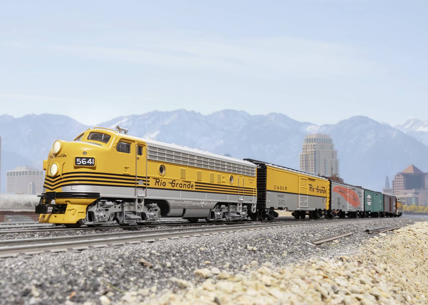 Train miniature jaune de la 'Rio Grande' circulant sur des rails, avec des montagnes et une ville en arrière-plan.