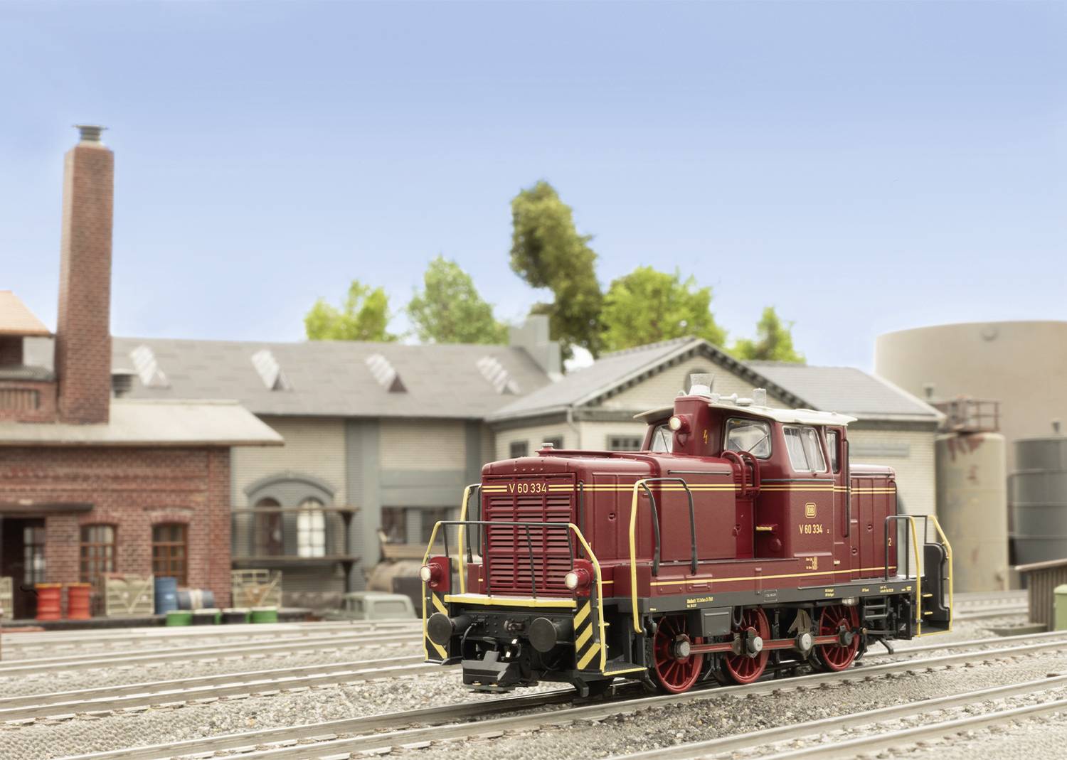 Un modèle de locomotive à vapeur rouge est posé sur un rail devant un bâtiment industriel miniature doté d'une cheminée et d'un réservoir d'eau.