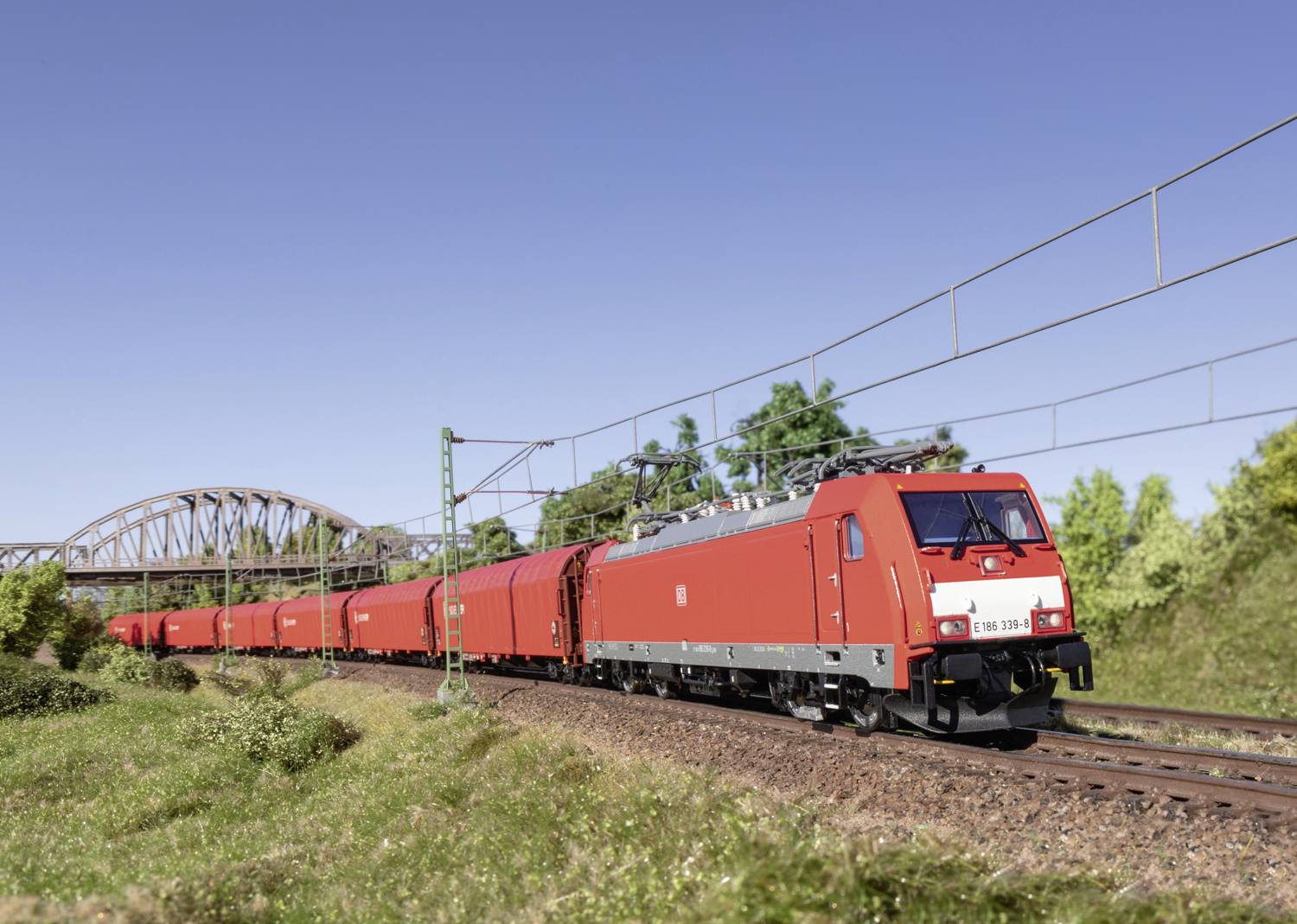 Un train de marchandises rouge circule de jour sur une voie ferrée à travers une région rurale. Un pont est visible en arrière-plan.
