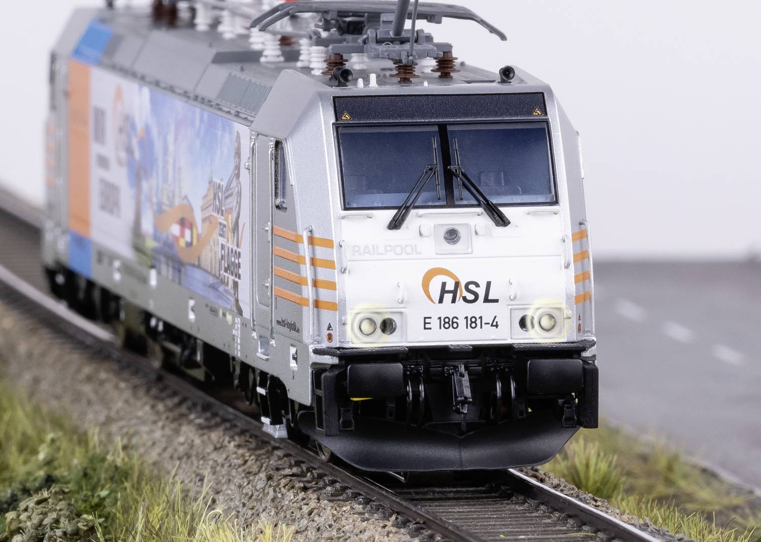 Un train miniature à l'échelle 1:87 circule sur un réseau de voies ferrées miniatures. La locomotive arbore le logo 'HSL' sur sa façade avant.