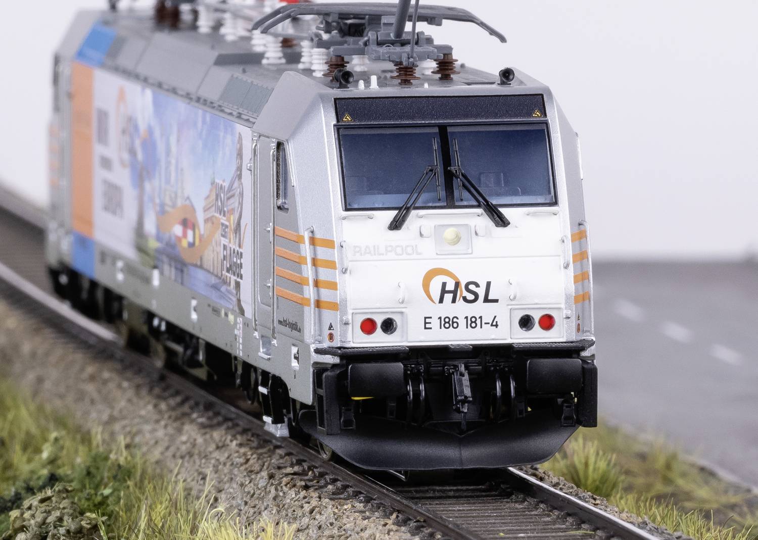 Une locomotive miniature sur une section de voie ferrée, marquée 'HSL' et portant le numéro 'E 186 181-4'. Elle est argentée avec un design coloré.