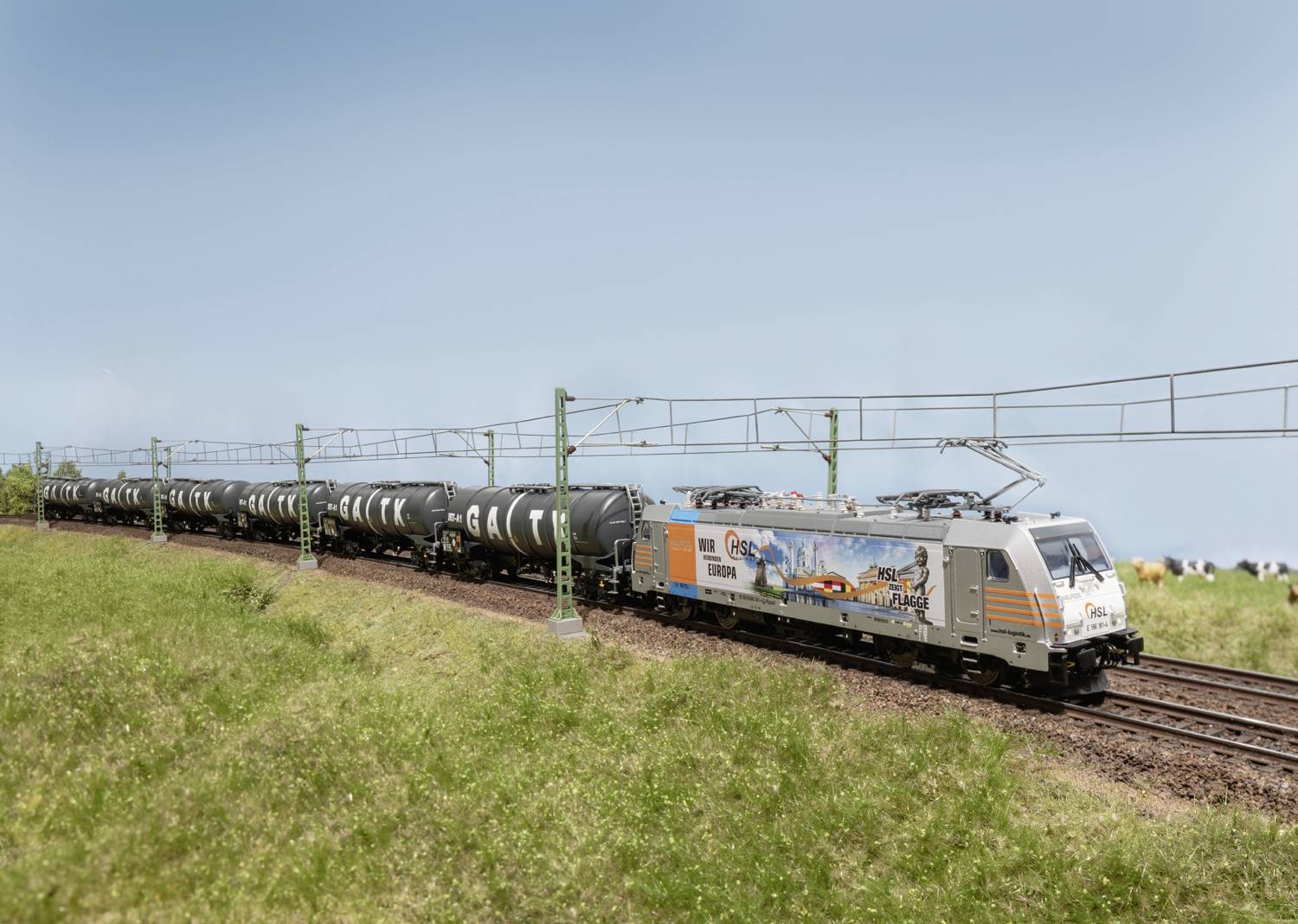 Un train de marchandises avec une locomotive peinte tire des wagons-citernes noirs à travers un paysage vert avec des lignes aériennes sous un ciel bleu.