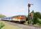 Un train orange et bleu circule sur une voie ferrée à travers un paysage verdoyant. Un signal mécanique est à l'arrêt.