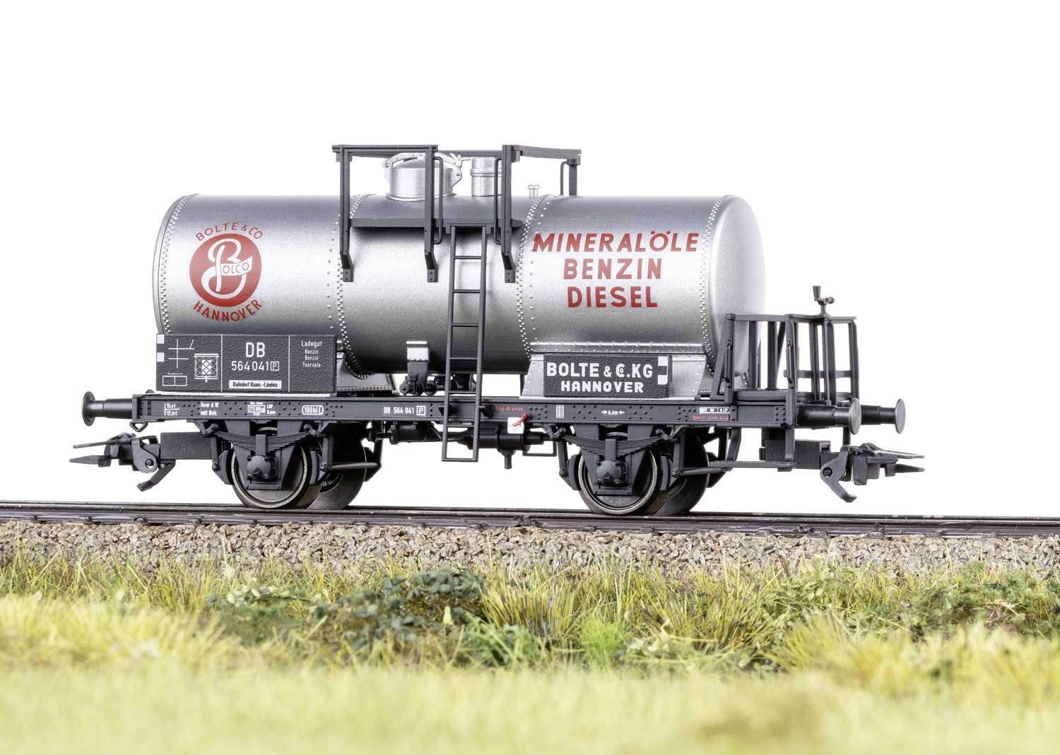 Un wagon-citerne argenté portant l'inscription 'Huiles minérales Essence Diesel', stationné sur des rails, entouré d'herbe.