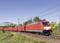 Un train de marchandises rouge circule sous un ciel clair à travers un paysage verdoyant et passe devant un pont ferroviaire en arrière-plan.