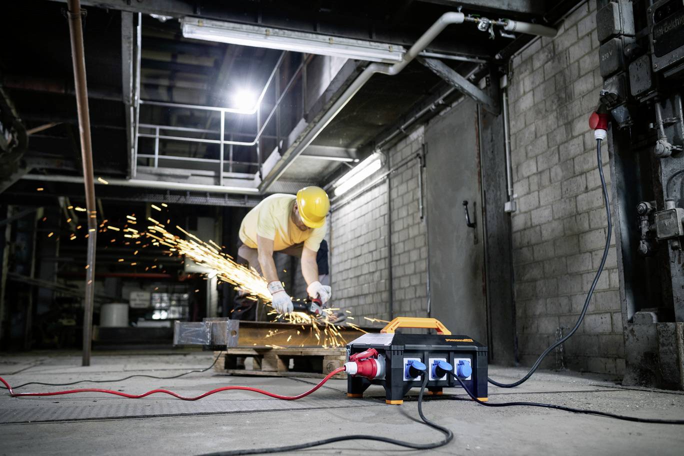 Un ouvrier portant un casque de sécurité meule du métal dans un hall industriel, tandis que des étincelles volent. Au premier plan, un coffret de distribution.