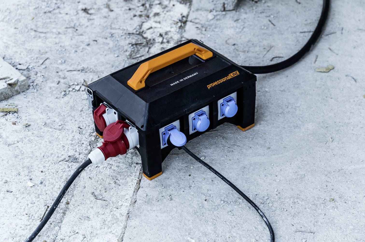 Un boîtier de distribution électrique noir avec quatre prises bleues et une fiche rouge est posé sur une surface en béton, relié à un câble.