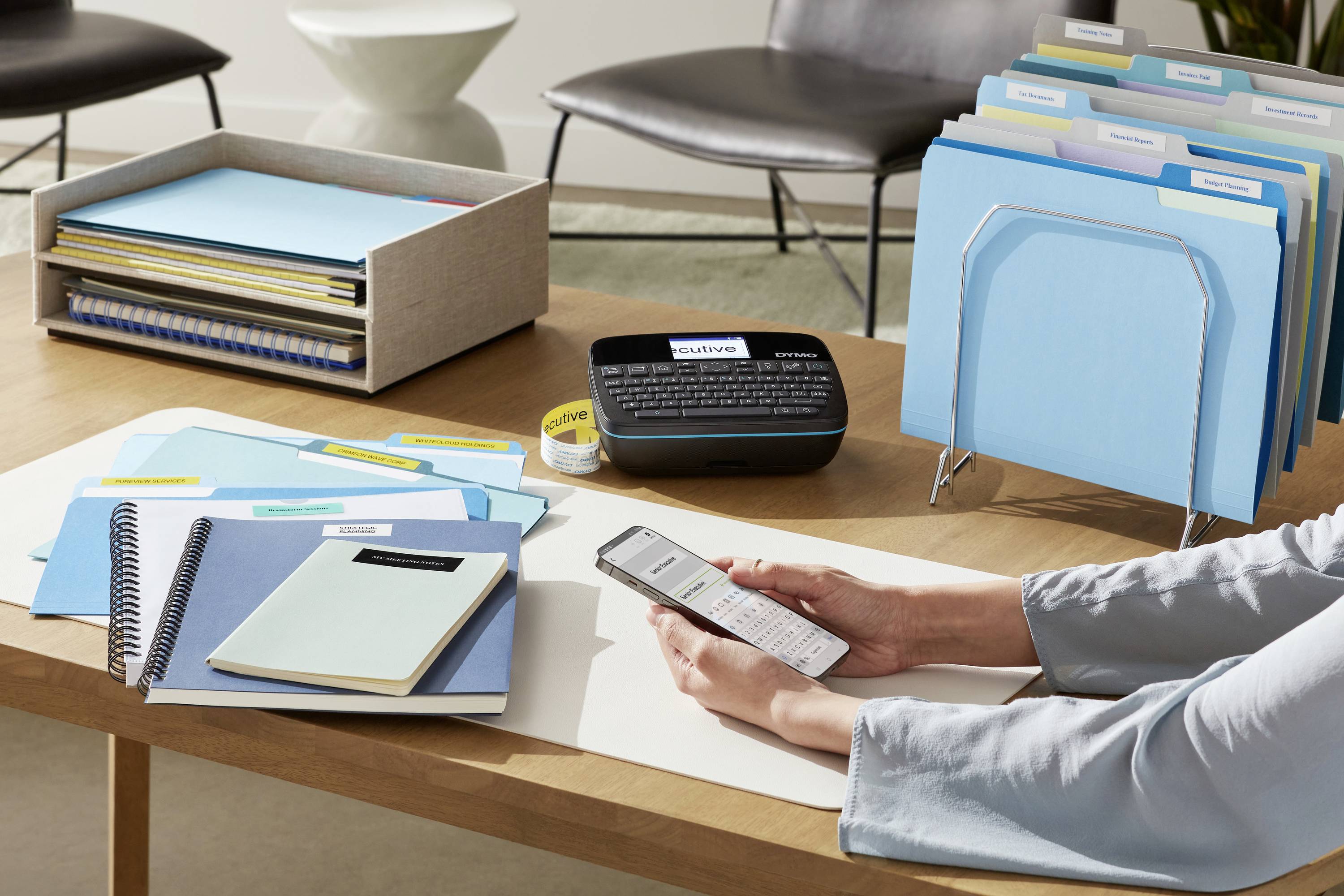 Une personne est assise à un bureau et utilise un appareil d'étiquetage. Des classeurs bleus et divers articles de bureau sont disposés sur la table.
