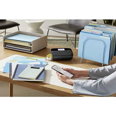 Une personne est assise à un bureau et utilise un appareil d'étiquetage. Des classeurs bleus et divers articles de bureau sont disposés sur la table.