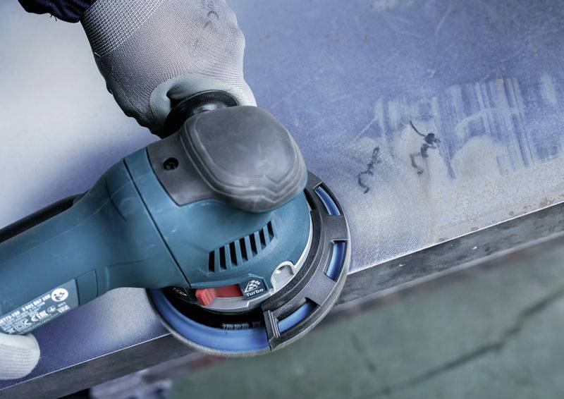 Une personne polit une surface métallique avec un meuleuse d'angle électrique. La machine est bleue avec une poignée noire.