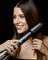 Une femme lisse ses longs cheveux bruns avec un fer à lisser noir sur un fond noir. Elle sourit, satisfaite.