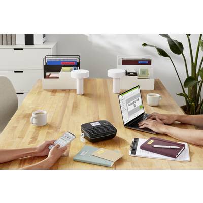 Sur une table, deux personnes travaillent avec une imprimante d'étiquettes et un ordinateur portable. Du matériel de bureau et des plantes sont en arrière-plan.
