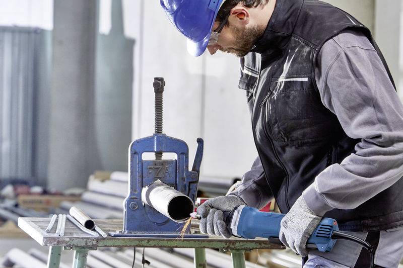 Un ouvrier portant des lunettes de protection bleues et un casque coupe un tuyau avec une scie dans un atelier.