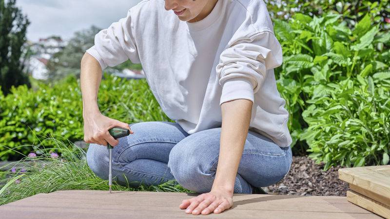 Une personne en tenue décontractée enfonce une vis dans une planche de bois à l'aide d'un tournevis, dehors dans un jardin.