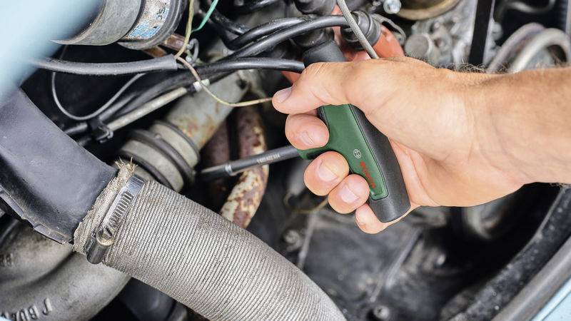 'Gros plan d'une main utilisant un tournevis pour travailler sur une pièce automobile. De nombreux câbles et tuyaux sont visibles.'